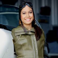 A woman smiling, wearing a green jacket and aviation goggles on her head, stands next to an aircraft, embodying the adventurous spirit found in social emotional learning curriculum.