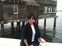 A person sits on a bench near a tranquil body of water, wearing sunglasses and a dark jacket over a white shirt. Behind them is a rustic wooden building on stilts. This scene evokes the calm reflection often found in SEL activities that encourage personal growth and connection.