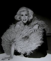 A black-and-white photo of a person with light hair, wearing a glamorous outfit adorned with large, feathery textures. They are smiling and posed against a dark background, embodying the elegance and variety celebrated in diversity programs.