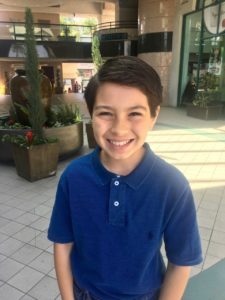 A smiling young boy with dark hair wears a blue polo shirt. He stands indoors in a brightly lit area adorned with potted plants and modern architecture, engaging students in get-to-know-you activities as part of their social-emotional learning curriculum.