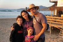 A family of four stands on a beach at sunset, embracing amidst mountains and the ocean. The father, in a straw hat, exudes warmth, embodying the joy often found in SEL activities that promote emotional connections. Smiles all around reflect a harmony celebrated through shared moments.
