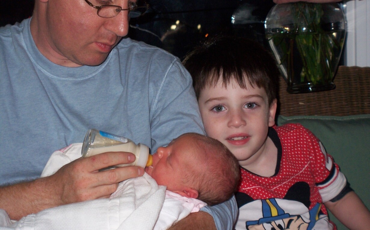A man bottle-feeding a baby while a young child in a Mickey Mouse shirt sits nearby, smiling at the camera, creates a warm scene of connection and nurturing. They're indoors, with a glimpse of flowers in the background—echoing themes from an SEL curriculum.