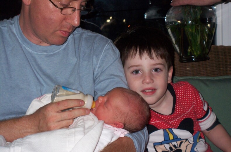 A man bottle-feeding a baby while a young child in a Mickey Mouse shirt sits nearby, smiling at the camera, creates a warm scene of connection and nurturing. They're indoors, with a glimpse of flowers in the background—echoing themes from an SEL curriculum.
