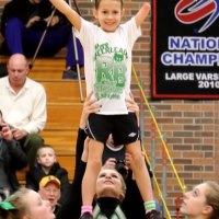 A young cheerleader in a white and green t-shirt is lifted by a team during a performance in a gym. Spectators, celebrating the spirit of diversity programs, are seated in the background near a banner that reads "National Champions.