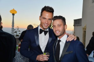 Two men in formal attire, both wearing dark suits and bow ties, stand close together at an outdoor event celebrating diversity programs. One holds a drink. The background shows a sunset with a lamp post and other people nearby.