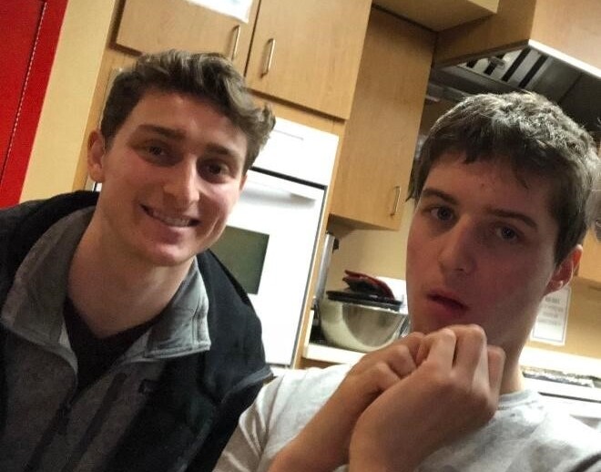 Two young men are in a kitchen. One smiles at the camera while the other looks off to the side, perhaps pondering their next step in social emotional learning activities. They stand before wooden cabinets and a countertop with a mixing bowl, blending ingredients of both recipes and relationships.