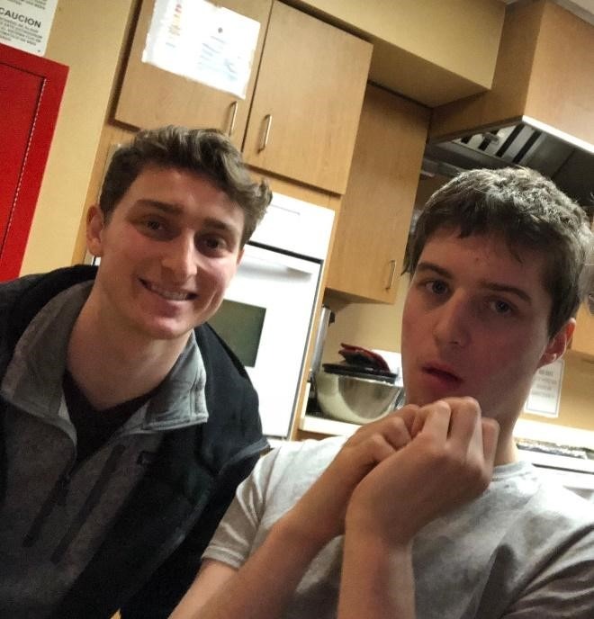 Two young men are in a kitchen. One smiles at the camera while the other looks off to the side, perhaps pondering their next step in social emotional learning activities. They stand before wooden cabinets and a countertop with a mixing bowl, blending ingredients of both recipes and relationships.