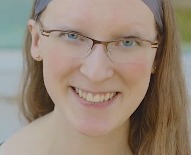 A person with long hair, wearing glasses and a gray headband, smiles at the camera. Dressed in a black patterned top, they exude warmth and confidence akin to an SEL curriculum leader, standing before a softly blurred background.