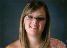 A person with long, light brown hair and glasses smiles at the camera, embodying the spirit of social emotional learning activities. Wearing a pink top against a dark, plain background, they exude warmth and openness.