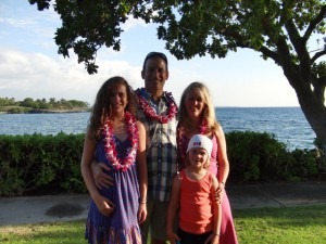 A family of four, wearing leis, stands together in front of the ocean. Two adults and two children pose under a tree, embraced by greenery and a clear sky. This bright and cheerful scene reflects the essence of social emotional learning activities, fostering connection and joy.