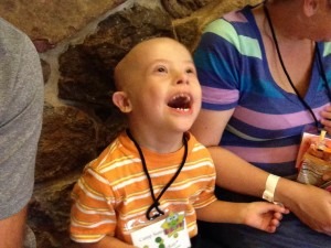 A child in an orange-striped shirt and a badge laughs joyfully, embodying the spirit of social emotional learning activities, as they sit between two adults—one in a gray shirt and the other in colorful stripes. The warm scene unfolds against a backdrop of a stone wall.