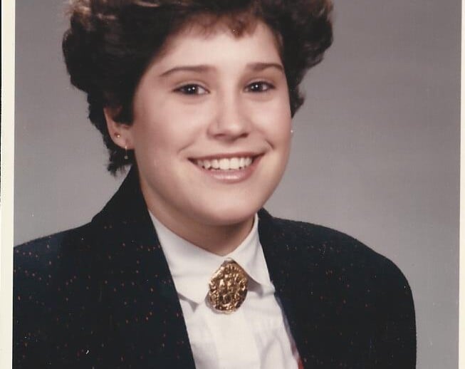 A person with short, curly hair is smiling at the camera, embodying the essence of SEL activities. They are wearing a dark blazer with a white shirt and a red vest, complemented by a decorative brooch that symbolizes their commitment to social emotional learning.