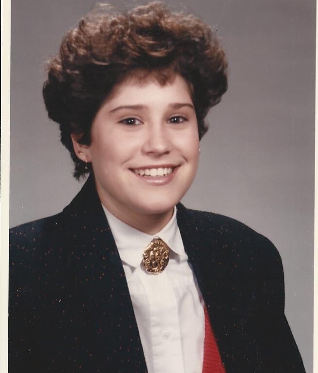 A person with short, curly hair is smiling at the camera, embodying the essence of SEL activities. They are wearing a dark blazer with a white shirt and a red vest, complemented by a decorative brooch that symbolizes their commitment to social emotional learning.