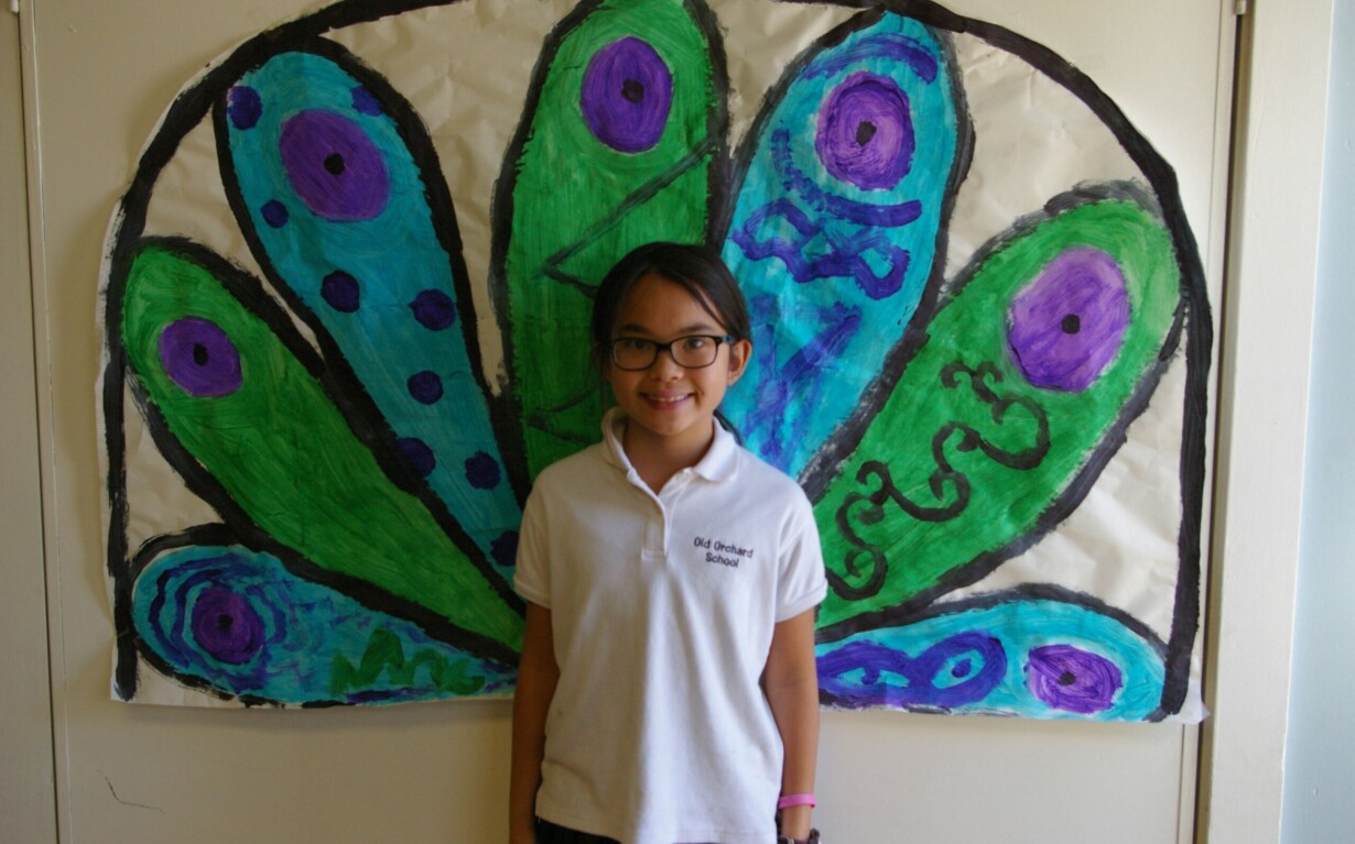 A child wearing glasses and a white shirt stands smiling in front of a colorful, hand-painted mural of peacock feathers in shades of green, blue, and purple—an inspiring backdrop for social emotional learning curriculum that celebrates creativity and self-expression.