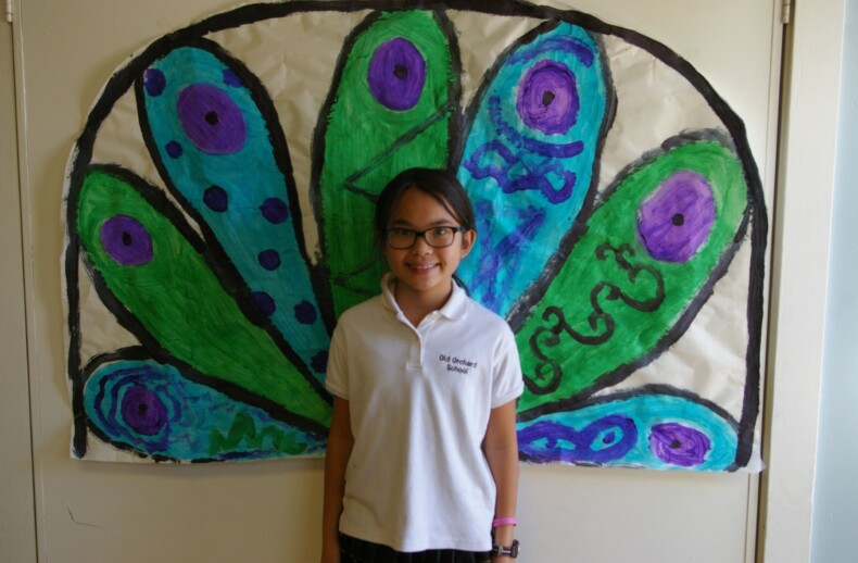 A child wearing glasses and a white shirt stands smiling in front of a colorful, hand-painted mural of peacock feathers in shades of green, blue, and purple—an inspiring backdrop for social emotional learning curriculum that celebrates creativity and self-expression.