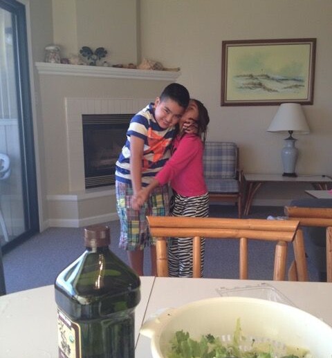 In a cozy living room, two children engage in a heartwarming social emotional learning activity. A boy in a striped shirt and plaid shorts embraces a girl in a pink top and striped pants. In the foreground, there's a table with salad and behind them is a fireplace and painting.