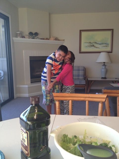 In a cozy living room, two children engage in a heartwarming social emotional learning activity. A boy in a striped shirt and plaid shorts embraces a girl in a pink top and striped pants. In the foreground, there's a table with salad and behind them is a fireplace and painting.