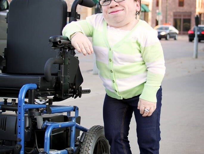 A person wearing a light green and white cardigan, jeans, and sandals leans against a motorized wheelchair on a sidewalk, with buildings in the background, embodying the essence of embracing diversity programs in everyday life.