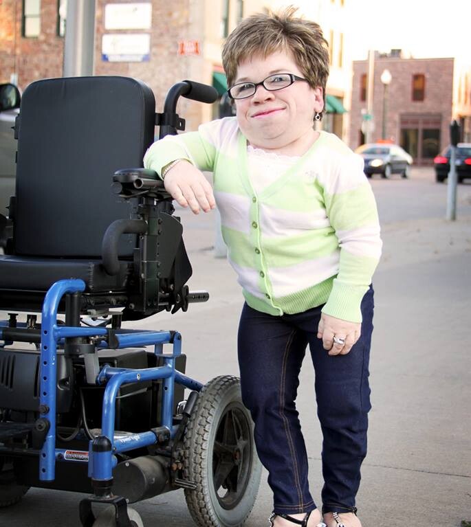 A person wearing a light green and white cardigan, jeans, and sandals leans against a motorized wheelchair on a sidewalk, with buildings in the background, embodying the essence of embracing diversity programs in everyday life.