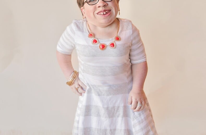 A person wearing glasses and a striped white dress stands on a soft carpet, exuding warmth akin to SEL activities. They are smiling, donning a statement necklace and black shoes. The background is a plain light color.