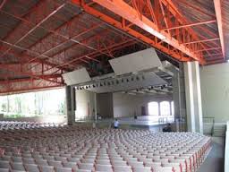 A spacious indoor auditorium with an empty stage and rows of white seats hosts social emotional learning activities. The ceiling features exposed red beams, and natural light filters in through large windows on the side, creating a calming ambiance for participants.