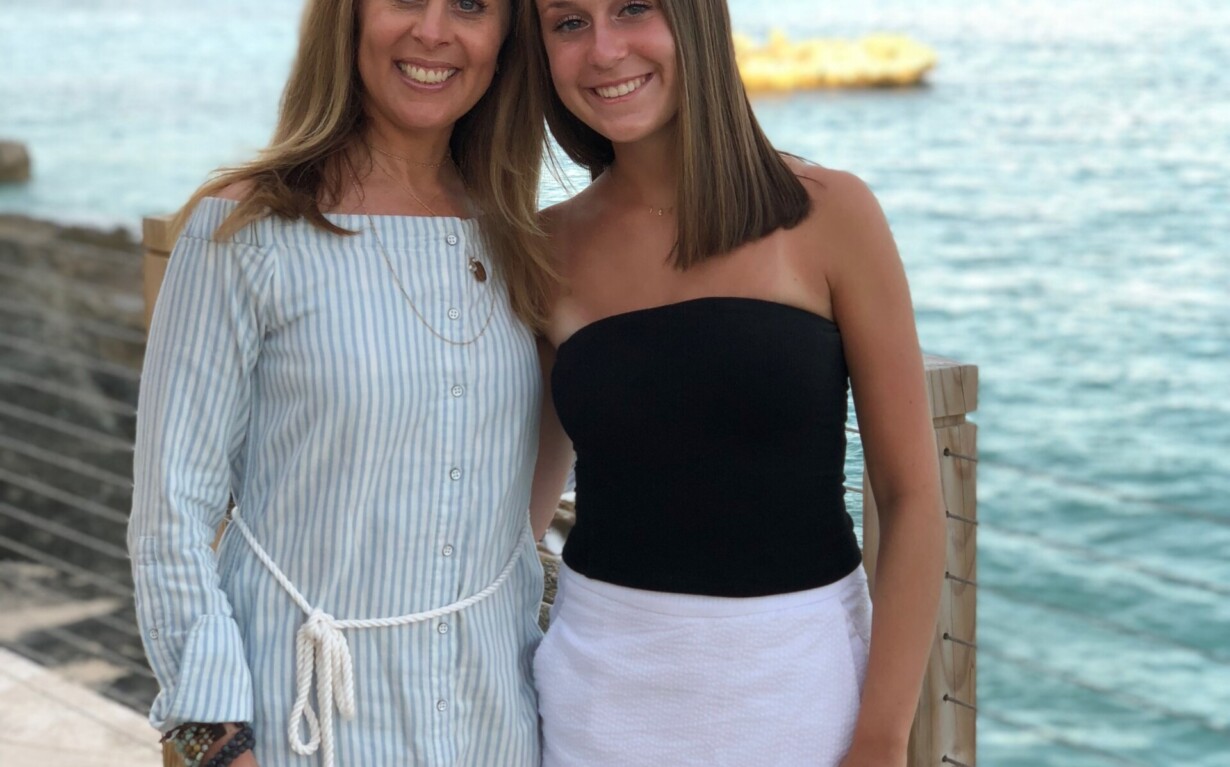 Two women are smiling and standing side by side near the ocean, embracing a spirit of unity akin to diversity programs. One wears a light blue, striped dress, and the other is in a black top with a white skirt. The background showcases calm blue water and a partly cloudy sky.