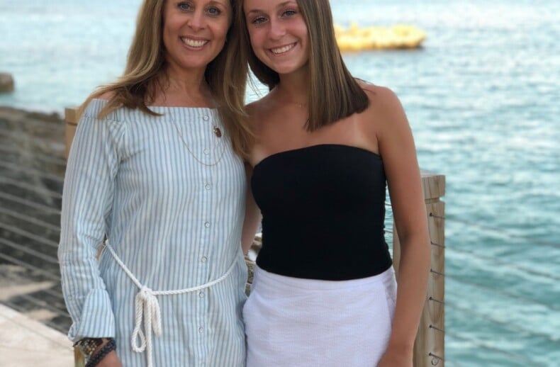 Two women are smiling and standing side by side near the ocean, embracing a spirit of unity akin to diversity programs. One wears a light blue, striped dress, and the other is in a black top with a white skirt. The background showcases calm blue water and a partly cloudy sky.