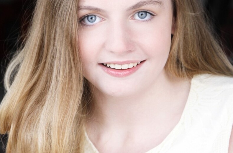 A person with long, blonde hair and blue eyes is smiling softly. Wearing a white top, they look directly at the camera. The blurred background highlights their face, much like how SEL activities focus on individual expression and emotional clarity.