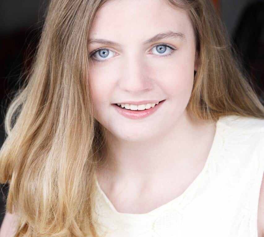 A person with long, blonde hair and blue eyes is smiling softly. Wearing a white top, they look directly at the camera. The blurred background highlights their face, much like how SEL activities focus on individual expression and emotional clarity.
