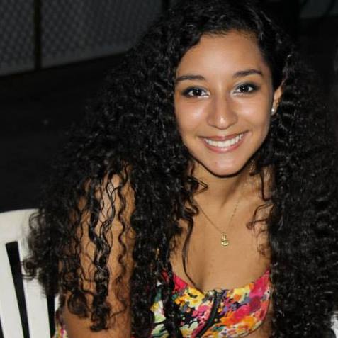 A person with long, curly hair smiles at the camera, embodying the warmth often fostered in diversity programs. They wear a floral-patterned top and a gold necklace with a pendant while seated on a white chair in an inviting outdoor setting.