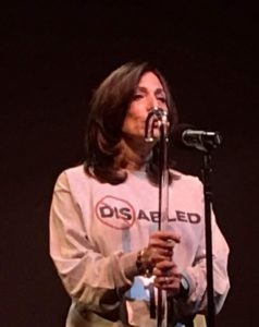 A woman stands on stage holding a microphone, wearing a sweatshirt with "DISABLED," where "DIS" is crossed out to emphasize "ABLED." Her performance inspires like social emotional learning activities, encouraging the audience to reflect on their own strengths in this intimate setting.