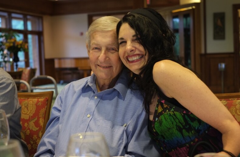 A smiling older man in a blue shirt sits next to a younger woman with long black hair in a colorful dress, embodying the essence of diversity programs. They are indoors, framed by wooden walls and a window, as the woman leans affectionately towards him.