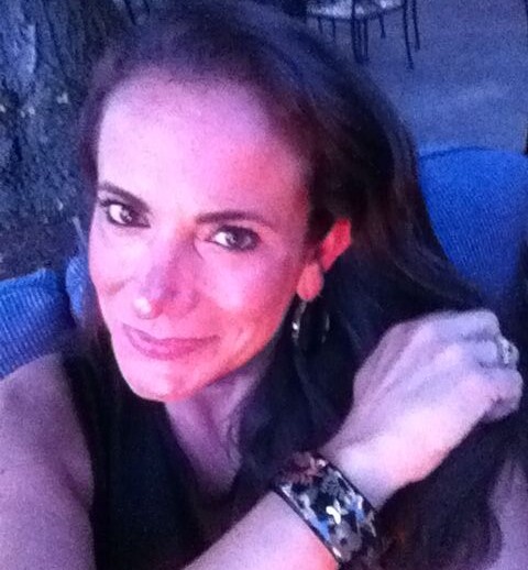 A woman with long brown hair is smiling at the camera, seated outdoors near a tree. She is wearing a bracelet and a dark sleeveless top, with chairs and tables visible in the background under evening light, reflecting the warmth of diversity programs that celebrate connection and joy.