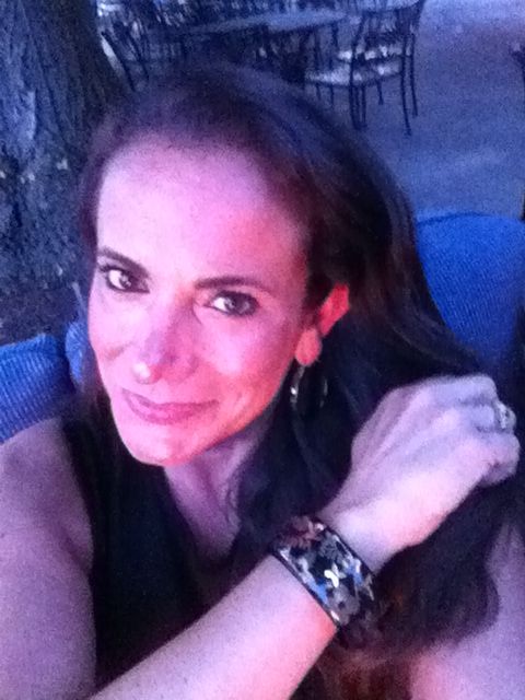 A woman with long brown hair is smiling at the camera, seated outdoors near a tree. She is wearing a bracelet and a dark sleeveless top, with chairs and tables visible in the background under evening light, reflecting the warmth of diversity programs that celebrate connection and joy.