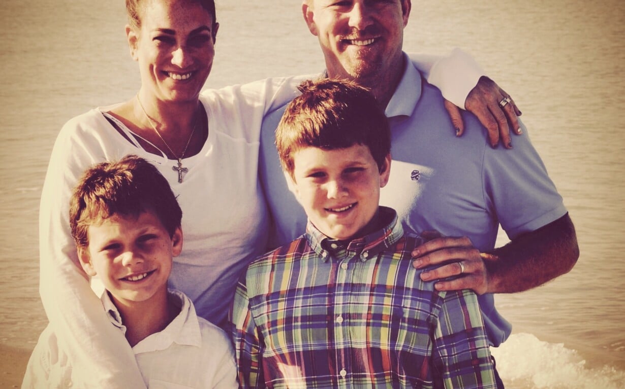 A family of four poses on the beach. The parents, a woman in a white shirt and a man in light blue, smile warmly behind their two young boys in plaid and white shirts. The ocean's gentle waves mirror the harmony of social emotional learning, fostering connection and joy.