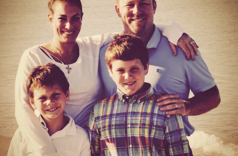 A family of four poses on the beach. The parents, a woman in a white shirt and a man in light blue, smile warmly behind their two young boys in plaid and white shirts. The ocean's gentle waves mirror the harmony of social emotional learning, fostering connection and joy.
