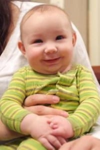 A smiling baby in a green striped outfit is being held, embodying the joy and comfort found in social emotional learning activities. With rosy cheeks and a content expression, the baby looks slightly to the side, reflecting a cozy sense of well-being.