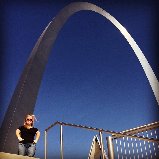 A person stands on a platform in front of the Gateway Arch against a clear blue sky, much like participants engaging in SEL activities that encourage them to reach new heights. The iconic stainless steel structure curves gracefully overhead.
