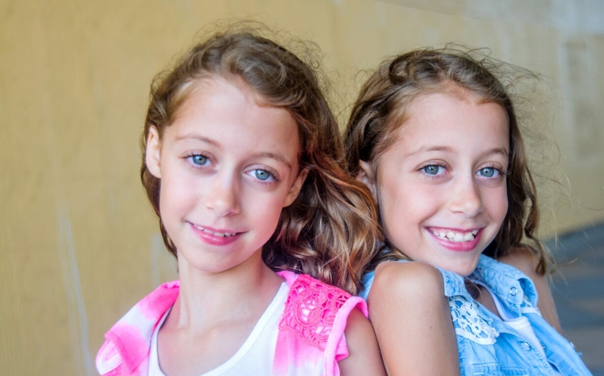 Two smiling young girls with long light brown hair stand closely together in front of a yellow background, embodying the spirit of diversity programs. One wears a pink top, and the other a blue top. With their light blue eyes, they appear cheerful and full of joy.