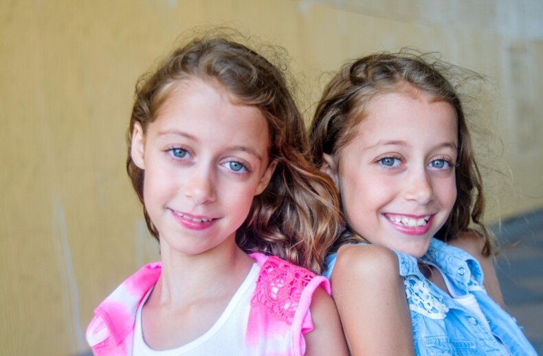 Two smiling young girls with long light brown hair stand closely together in front of a yellow background, embodying the spirit of diversity programs. One wears a pink top, and the other a blue top. With their light blue eyes, they appear cheerful and full of joy.