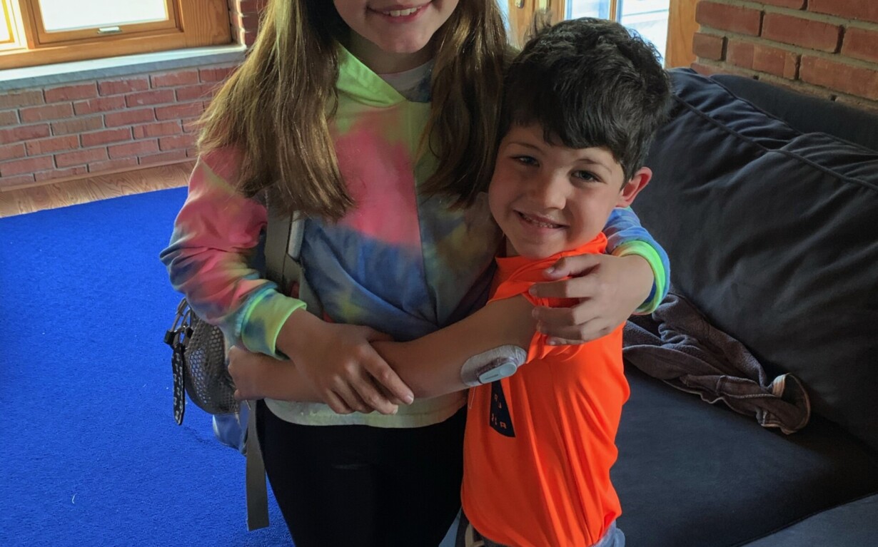 A girl in a tie-dye hoodie and a boy in an orange shirt embrace warmly inside a room with brick walls and large windows. This heartfelt moment on the blue carpet beside the gray sofa echoes themes from diversity programs, fostering connection and understanding.