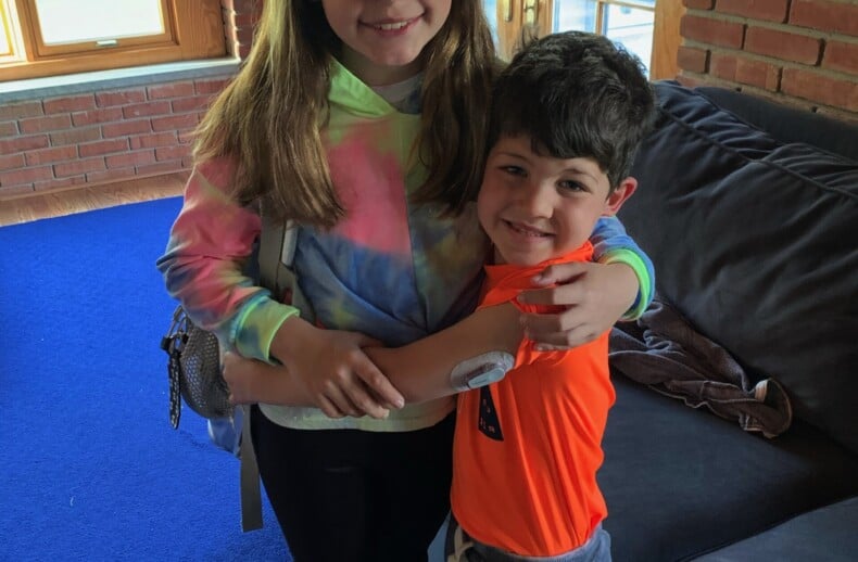 A girl in a tie-dye hoodie and a boy in an orange shirt embrace warmly inside a room with brick walls and large windows. This heartfelt moment on the blue carpet beside the gray sofa echoes themes from diversity programs, fostering connection and understanding.