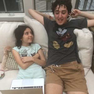 A young girl and a taller boy are sitting on a beige couch, engaging in an informal get-to-know-you activity. The girl, on the left with a laptop on her lap, gazes at the boy who is leaning back, smiling with hands behind his head. Both are dressed in casual clothes.