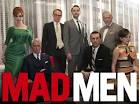 A diverse group of seven well-dressed individuals poses in front of window blinds, standing and sitting around two chairs. The words "Mad Men" are prominently displayed at the bottom in bold red and white letters, reminiscent of get-to-know-you activities where personalities boldly stand out.