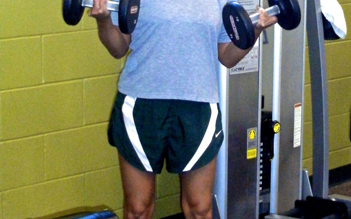 A person in a red cap, gray T-shirt, and green shorts is lifting dumbbells in a gym, integrating focus from their SEL curriculum. They stand confidently on a gym mat in front of exercise equipment.