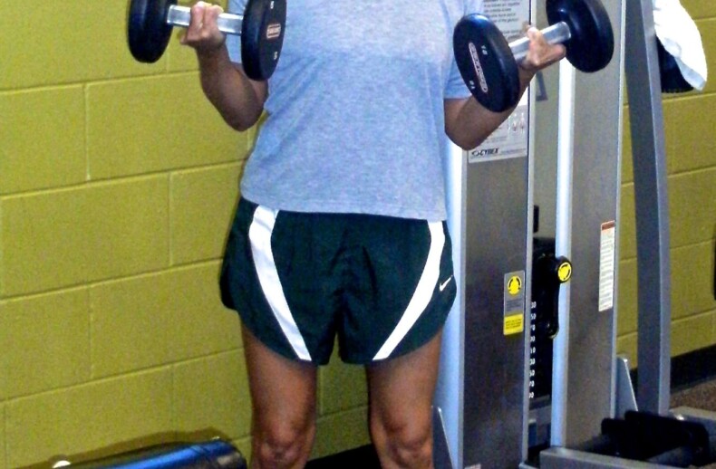 A person in a red cap, gray T-shirt, and green shorts is lifting dumbbells in a gym, integrating focus from their SEL curriculum. They stand confidently on a gym mat in front of exercise equipment.