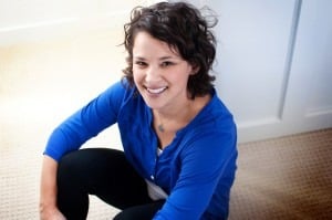 A person with curly hair, wearing a blue shirt, smiles while seated on a light-colored carpet near a white wall, embodying the warmth often found in social emotional learning curriculum settings.