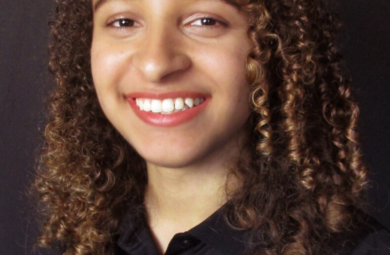 A woman with curly hair smiles warmly against a black background, wearing a black "PCCA Gibbs High School" polo shirt—a proud participant in diversity programs.