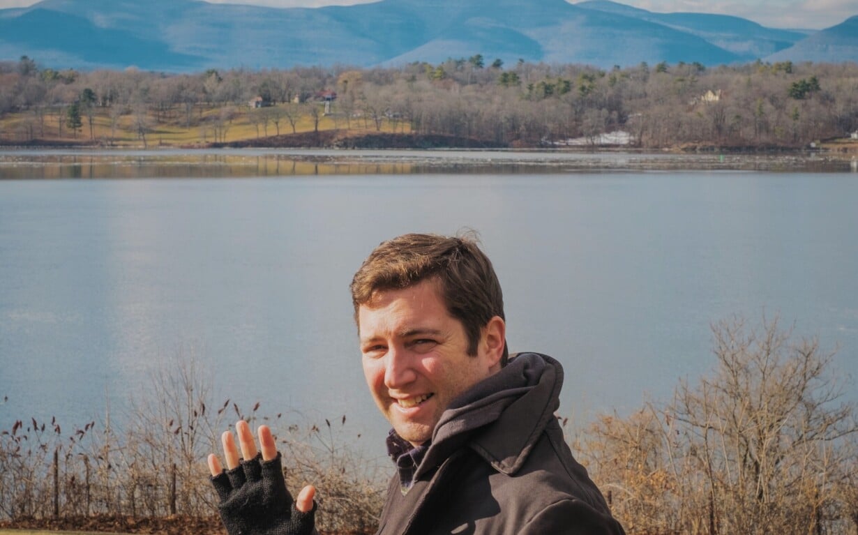A person wearing a dark coat and fingerless gloves is smiling and waving in front of a scenic lake, embodying the joy often found in SEL curriculum. The background features rolling hills and a partly cloudy sky, painting a picture of tranquility and reflection.