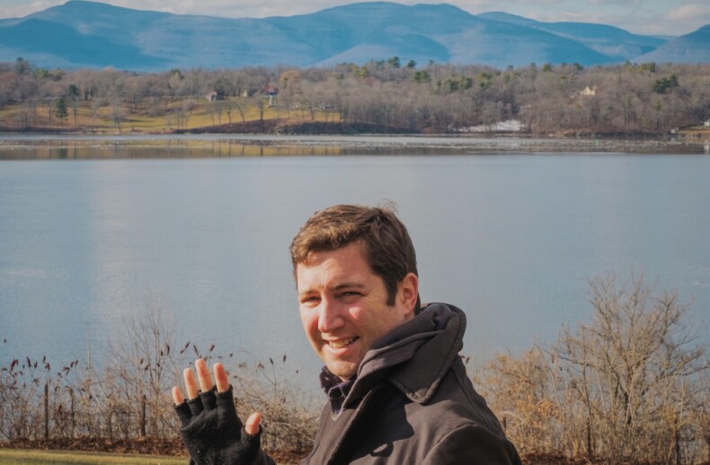 A person wearing a dark coat and fingerless gloves is smiling and waving in front of a scenic lake, embodying the joy often found in SEL curriculum. The background features rolling hills and a partly cloudy sky, painting a picture of tranquility and reflection.